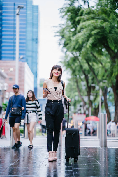 attractive Asian Chinese woman books a ride through her ride hailing app on her smartphone. She is walking with her luggage and is stylishly dressed in work wear  - Фото, изображение