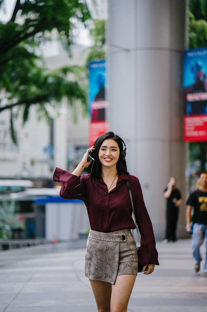 beautiful Korean woman in an elegant blouse and shorts dancing as she walks down a street in the city with her headphones on - Fotografie, Obrázek