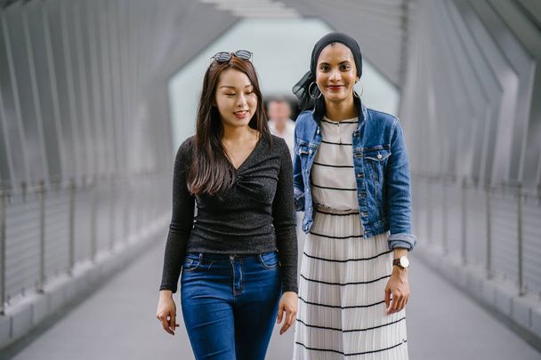 Two friends of different ethnicity on a bridge during the day. One is a Malay Muslim woman and the other is Chinese Asian. They are both young, energetic and attractive. - Foto, Imagem