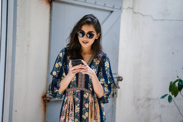 Portrait of a young, attractive and beautiful Indian Asian woman wearing a dress and sunglasses with her smartphone - Foto, Imagen