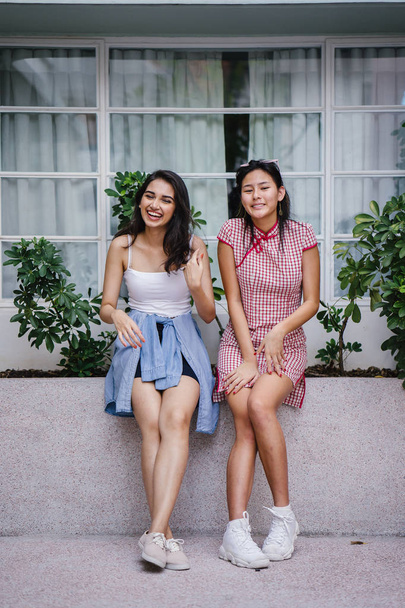 two young women friends of different ethnicity hanging out together. One is a Chinese woman in a cheongsam and the other an Indian woman in a trendy outfit - Photo, Image