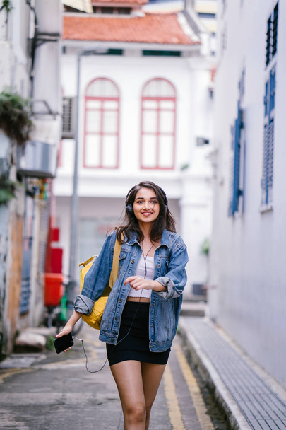 Portrait of a young and attractive Indian Asian woman in a denim jacket and yellow backpack enjoying music on her headphones - Фото, зображення