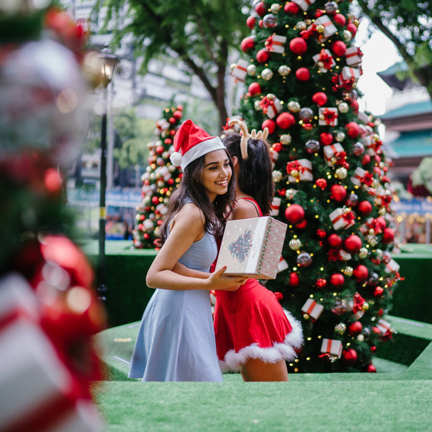 Two young Asian girls exchange presents with one another under a decorated Christmas tree in a street in the city during the day. One is Indian, the other Chinese - Valokuva, kuva
