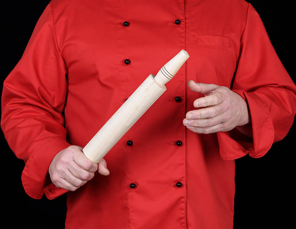 koken in een rood uniform houden een houten deegroller, zwarte achtergrond - Foto, afbeelding