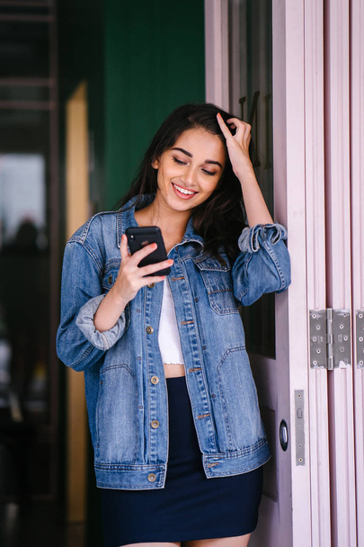Portrait of a beautiful, tall, elegant and young Indian Asian woman texting on her smartphone. She is wearing a retro denim jacket  - Foto, imagen