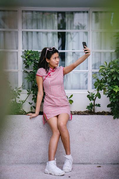Fashion portrait of a young, too-cool-for-school Asian Chinese teenager girl in a retro, traditional Chinese cheongsam dress taking a selfie of herself. - Φωτογραφία, εικόνα