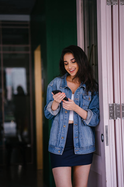 Portrait of a beautiful, tall, elegant and young Indian Asian woman texting on her smartphone. She is wearing a retro denim jacket  - Photo, image