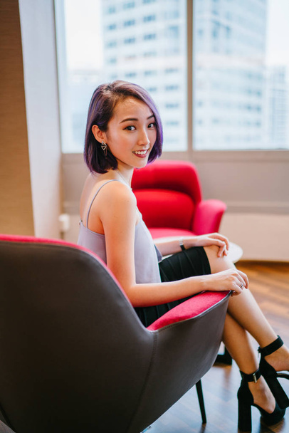 A young, slim and elegant Asian Chinese woman in a professional dress and dyed purple hair sits in a red armchair in her office coworking space. She is smiling candidly. - Фото, зображення