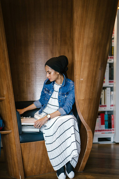 Giovane, alta, sottile, attraente e bella donna musulmana malese seduta in uno stand della biblioteca e lettura. Sta studiando e facendo ricerca da sola. - Foto, immagini