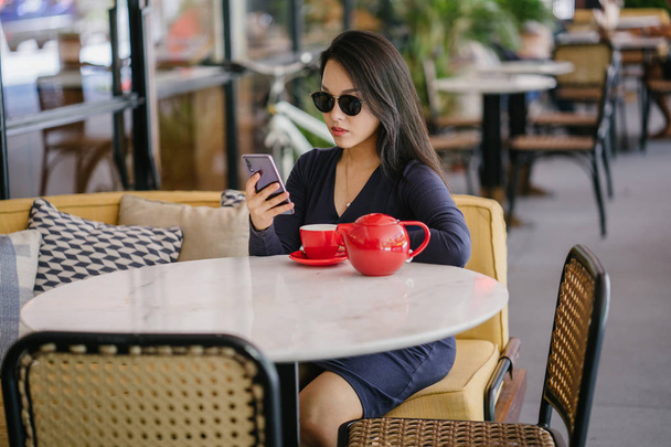 Πορτραίτο μιας νεαρής, ελκυστικής, κομψής και όμορφης ασιατικής γυναίκας που κάθεται έξω σε ένα υπαίθριο καφέ και απολαμβάνει ένα ζεστό φλιτζάνι τσάι από ένα σετ κόκκινου τσαγιού.  - Φωτογραφία, εικόνα