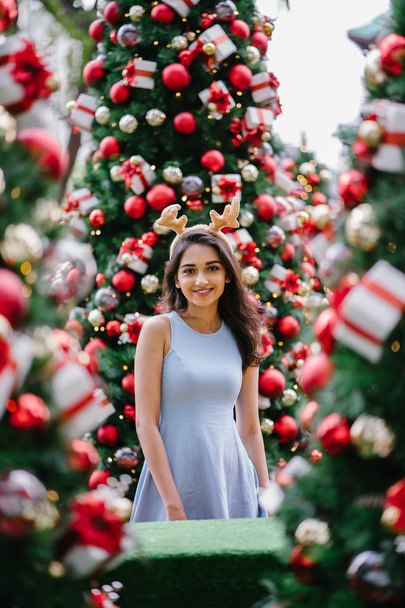Festive Christmas portrait of a young, beautiful and happy Indian Asian woman smiling as she holds a gift in front of a Christmas tree. - Foto, Imagem