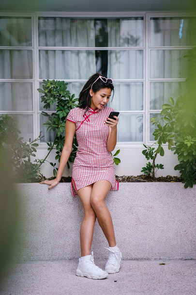 Fashion portrait of a young, too-cool-for-school Asian Chinese teenager girl in a retro red and white cheongsam dress sitting on a ledge during the day - Photo, image