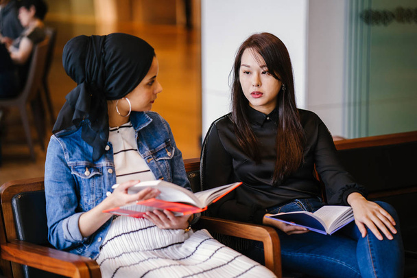 Two young female friends sitting on bench and reading books and discussing. One is a Muslim Malay woman and the other a Chinese woman.  - Foto, Imagem