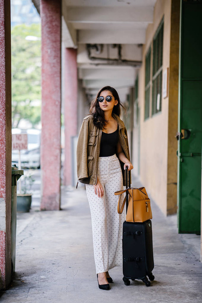 full-body portrait of a young, Indian Asian (Pubjabi) woman standing on the street with her luggage. She looks elegant, wealthy and stylish with her pant suit outfit and sunglasses - Foto, imagen