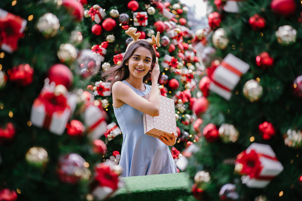 Festive Christmas portrait of a young, beautiful and happy Indian Asian woman smiling as she holds a gift in front of a Christmas tree. - Foto, Imagen