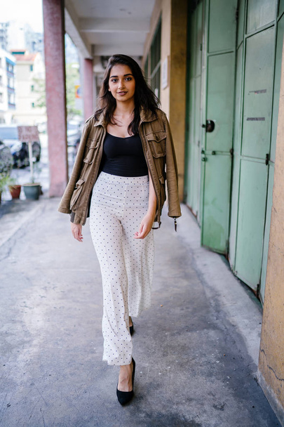 Portrait of a tall, beautiful, slim and elegant Indian Asian woman walking down a walkway in a city in Asia during the day. She is elegantly dressed and has a jacket draped over her shoulders. - Photo, image