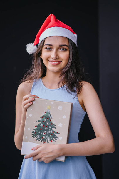 Portrait of a young and beautiful Indian Asian woman in a blue dress and red Santa hat against a black studio background. She is smiling happily as she holds onto a gift for a loved one. - Foto, Bild