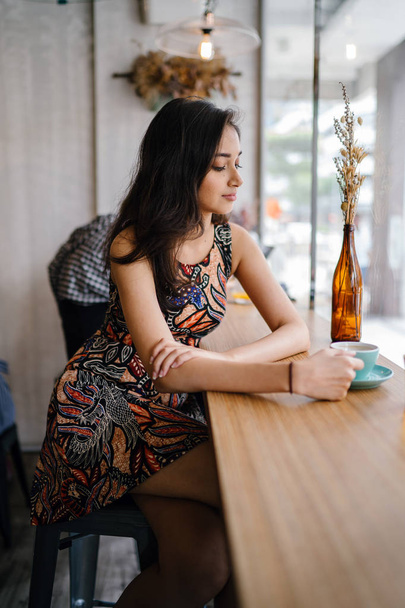 Young and beautiful Indian Asian Singaporean woman in a print dress sits in a cafe as she enjoys the weekend in Asia. - Foto, immagini