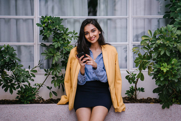 beautiful, young and stylish Indian Asian woman texting on her smartphone as she leans against a ledge in the day. She is wearing a retro yellow jacket over casual clothes - Photo, image
