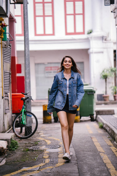 tall, young and elegant Indian Asian woman walking down an alley in the city during the day. She is wearing a retro denim jacket - Zdjęcie, obraz