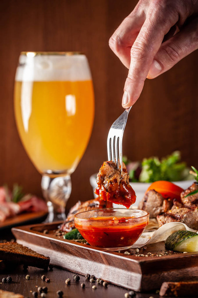 The concept of Georgian cuisine. A large wooden board with a kebab of beef, pork and chicken. Barbecue sauce and ketchup. In the background a glass of light beer with foam - Photo, image