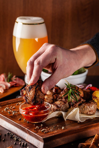 The concept of Georgian cuisine. A large wooden board with a kebab of beef, pork and chicken. Barbecue sauce and ketchup. In the background a glass of light beer with foam - Valokuva, kuva