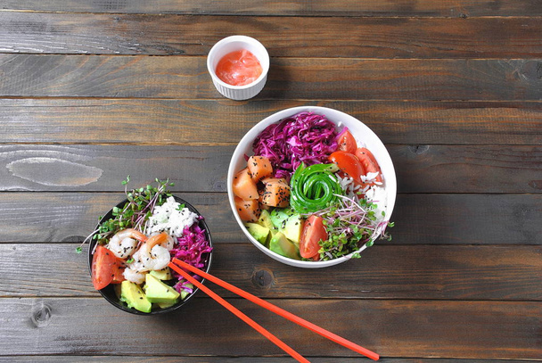 Organic food. Fresh seafood recipe. Two fresh poke bowls with salmon, shrimps, rice, red cabbage, avocado, cherry tomatoes and radish sprouts on wooden background. Food concept Poke Bowl  - Photo, Image