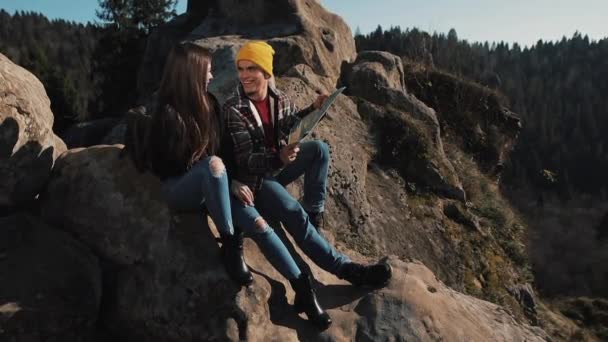 Travel to the mountains. Beautiful couple in love sits on the rock and learns a map. They talk with each other, laughing and having fun. - Materiaali, video