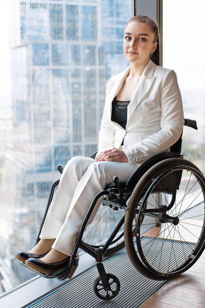 Confident happy businesswoman in wheelchair against the background of a panoramic window overlooking the skyscrapers and a big city., she is smiling at camera, disability overcoming concept - Photo, Image