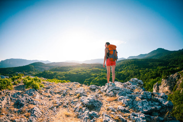 Girl at the top of the mountain. A woman with a backpack is standing on a rock. Climb to the top. Travel to picturesque places. Tourist against the sky and the sea. The brunette looks at the horizon. - Φωτογραφία, εικόνα