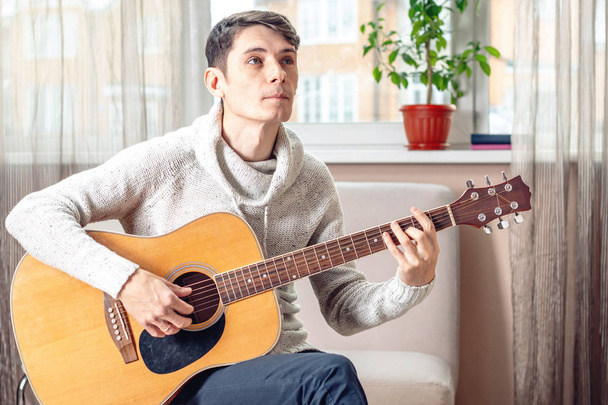 Young attractive male musician sitting on a chair playing acoustic guitar. The concept of music as a hobby - Foto, afbeelding