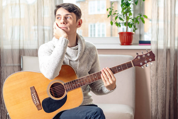 Νεαρός ελκυστικό αρσενικό μουσικός που κάθεται σε μια καρέκλα κρατώντας μια ακουστική κιθάρα. Η έννοια της μουσικής ως χόμπι - Φωτογραφία, εικόνα