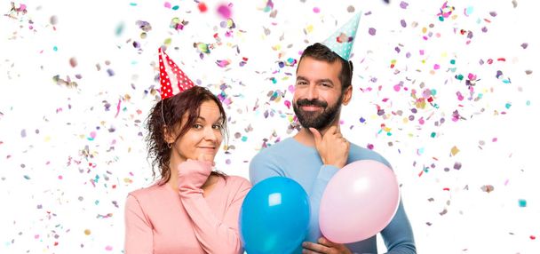 pareja con globos y sombreros de cumpleaños sonriendo y mirando al frente con cara confiada con confeti en una fiesta
 - Foto, Imagen
