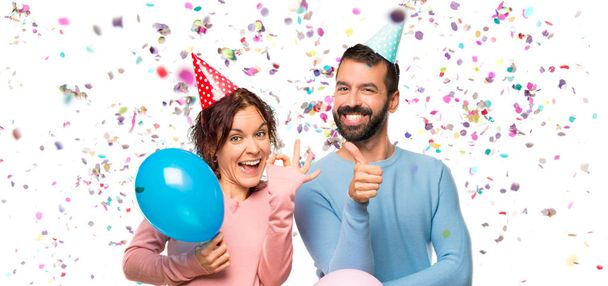 Paar mit Luftballons und Geburtstagsmützen zeigt ok Zeichen und gibt einen Daumen nach oben Geste mit der anderen Hand mit Konfetti in einer Party - Foto, Bild
