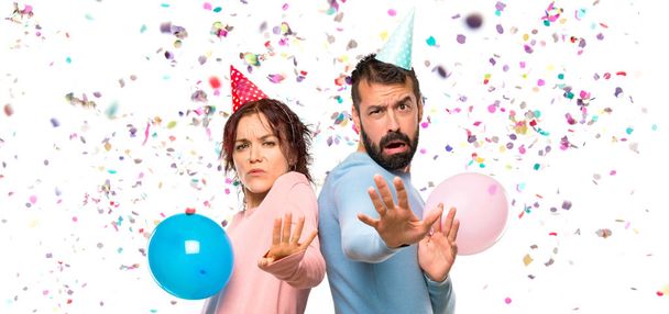 couple with balloons and birthday hats is a little bit nervous and scared stretching hands to the front with confetti in a party - Foto, immagini
