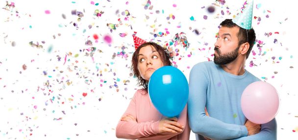 Paar mit Luftballons und Geburtstagsmützen macht unwichtige Geste, während es auf einer Party die Schultern mit Konfetti hebt - Foto, Bild