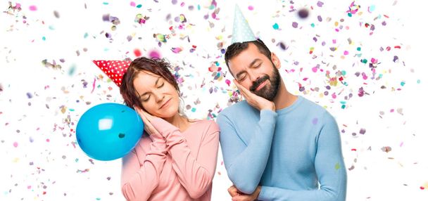 couple with balloons and birthday hats making sleep gesture. Adorable sweet expression with confetti in a party - Φωτογραφία, εικόνα