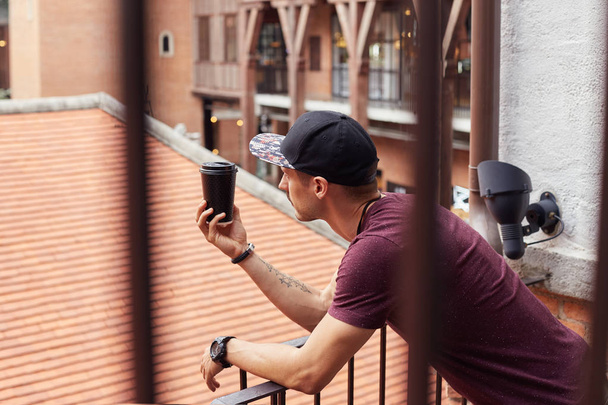 Portrait through stair rails of attractive young Caucasian man in stylish wear enjoying fresh takeaway coffee in paper cup, standing in urban setting over street background. People, lifestyle concept. - Foto, afbeelding