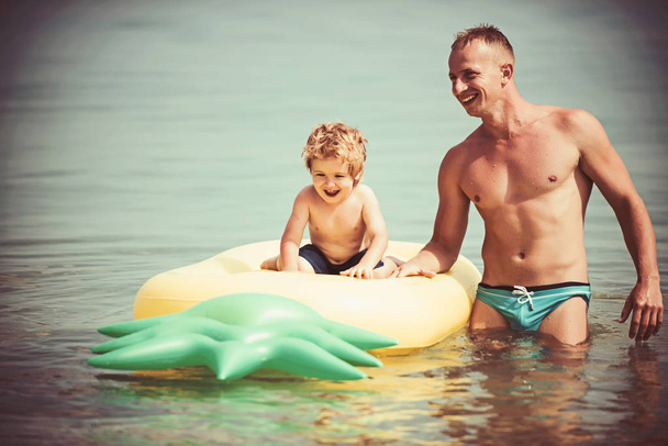 Maldives or Miami beach activity joy. Father and son swim in water on fathers day. Pineapple inflatable or air mattress. Happy family on Caribbean sea. Summer vacation and travel to ocean, vintage. - Photo, image