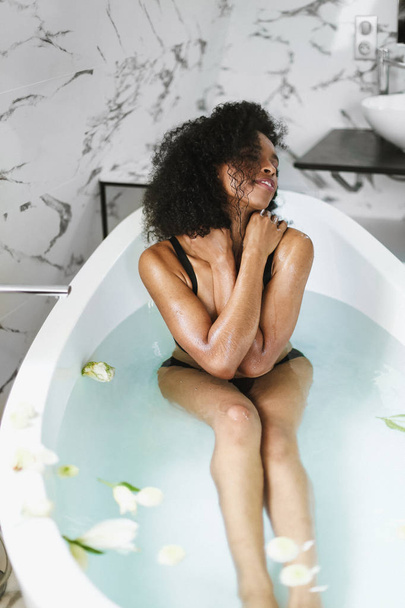Young happy afro american girl sitting in bath, wearing black swimsuit. - Foto, imagen