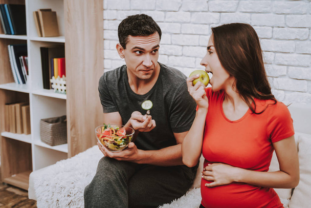 Pár ül a kanapén. Nő egy almát eszik. Nő egy fiatal terhes lány. Ember van Giving egy uborka saláta a nő. Saláta tál friss zöldség. Az emberek otthon található. - Fotó, kép