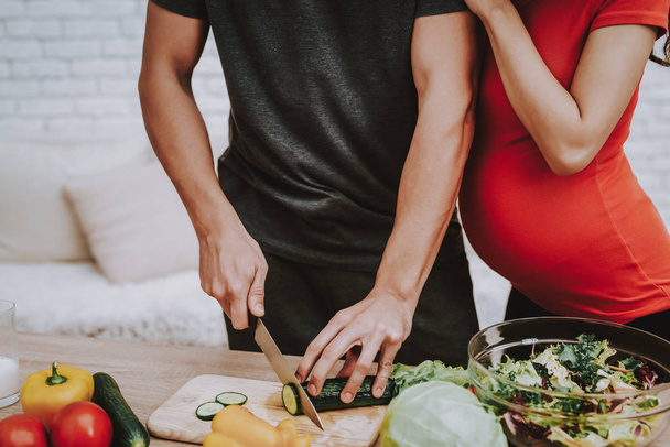 Man is Cooking Salad. Woman is Hugging Her Man. Woman is a Pregnant Girl. Man is Cutting the Cucumber a Knife. Vegetables, Milk and Salad in Bowl on Table. People Located at Home Interior. - Фото, изображение