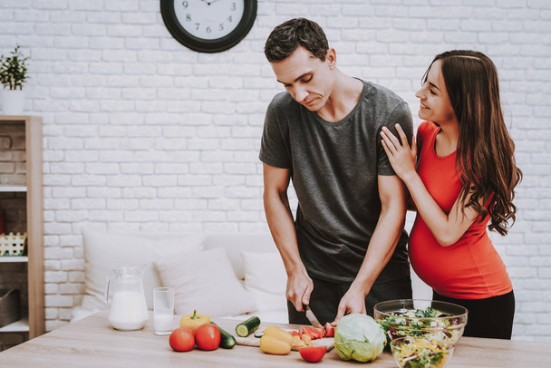 Man is Cooking Salad. Woman is Hugging Her Man. Woman is a Pregnant Girl. Man is Cutting the Tomatoe a Knife. Vegetables, Milk and Salad in Bowl on Table. Girl is Smiling. People Located at Home. - Фото, изображение