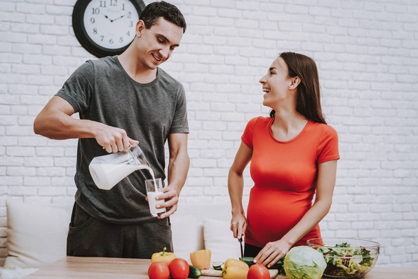 Woman is Cooking Salad. Woman is Pregnant Girl. Woman is Cutting the Cucumber a Knife. Man is Pouring a Milk in Cup from Jar. Vegetables and Salad in Bowl on Table. Persons is Smiling. People at Home. - 写真・画像