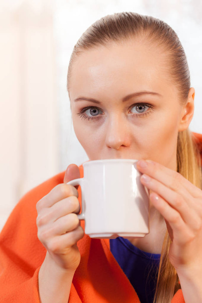 Young woman lying on sofa under warm blanket drinking tea, coffee or different hot drink from mug enjoying her leisure time during cold weather. - Foto, Bild