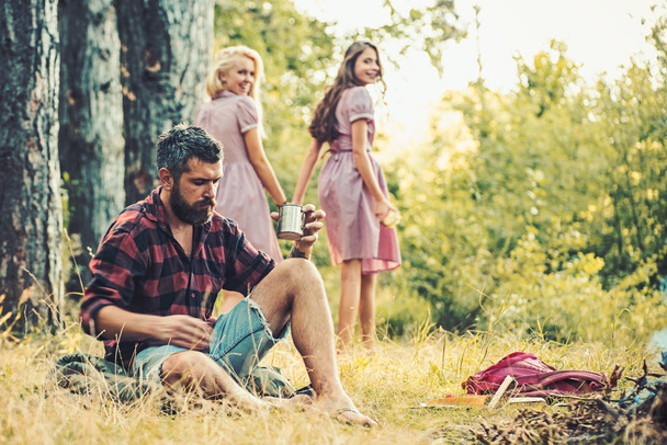 Friends enjoy camping in forest. Bearded man drink tea at bonfire with women on blurred background. Hipster with beard sit at campfire. Summer vacation concept. Camping and hiking and travel - Foto, Imagen