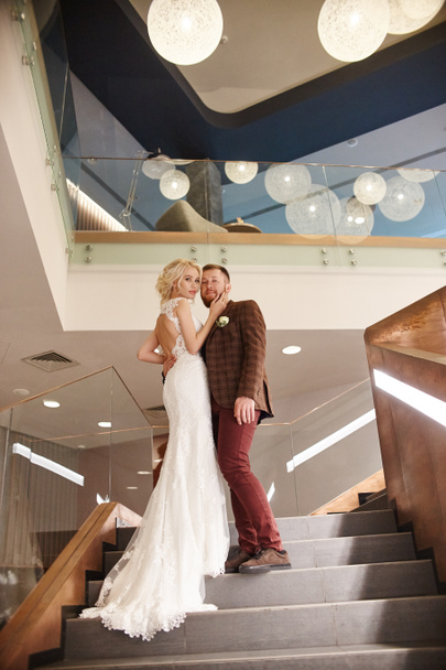 Νύφη σε ένα κομψό μακρύ φόρεμα με ένα τρένο και το γαμπρό σταθεί στα μεγάλα σκαλοπάτια, ζευγάρι στην αγάπη αγκαλιές στις σκάλες φιλί και βλέμμα στο άλλο. Την ημέρα του γάμου, ζευγάρι για την ημέρα της εγγραφής του γάμου - Φωτογραφία, εικόνα