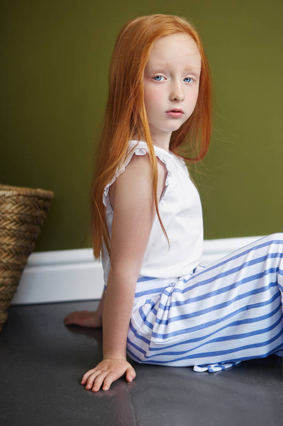 Little red-haired girl with a basket of flowers posing on an olive background. Spring portrait of a redhead girl with blue eyes. Hair the color of fire, Norwegian teen girl - Foto, Imagem