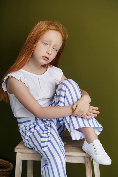 Little red-haired girl with a basket of flowers posing on an olive background. Spring portrait of a redhead girl with blue eyes. Hair the color of fire, Norwegian teen girl. - Photo, Image