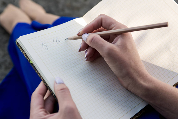girl in a blue dress sitting on the steps and writing a pencil in a notebook - Foto, Imagen